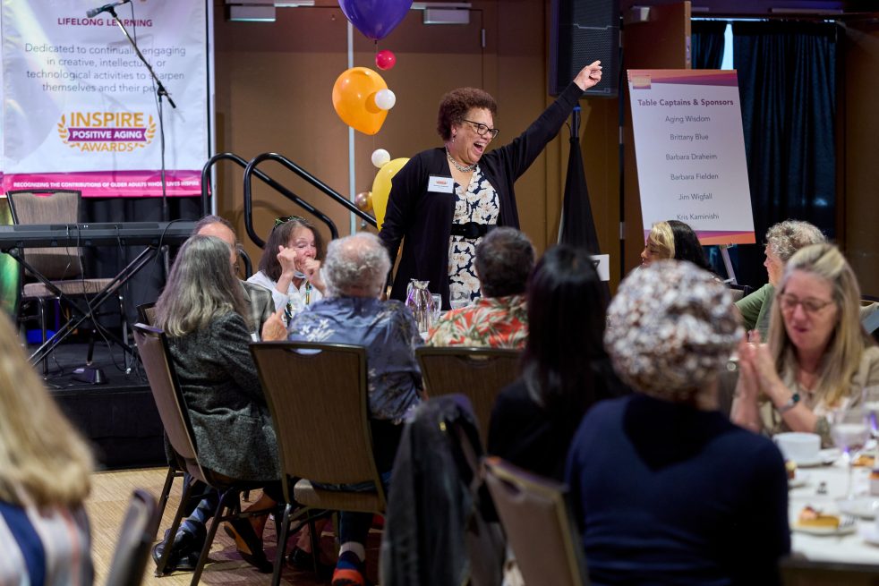 a woman stands and points one finger in the air as if dancing, in a crowded event room