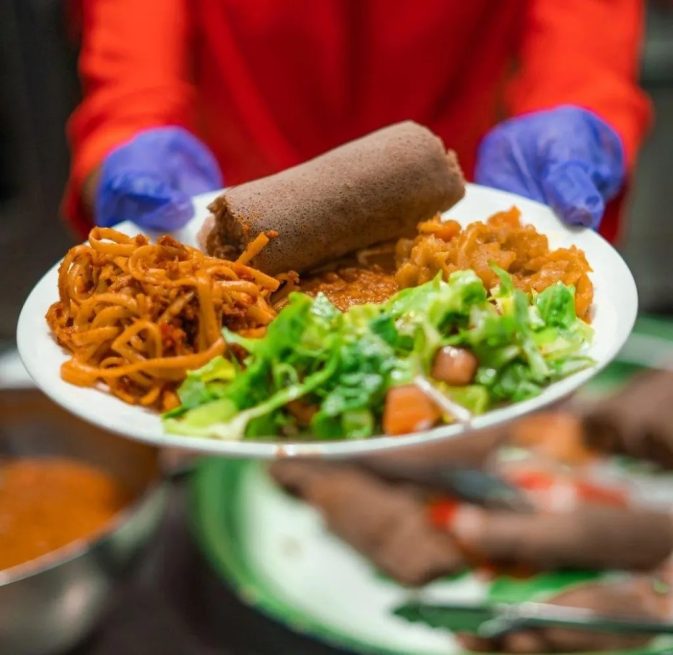 A plate of injera, salad, and noodles being handed toward the camera