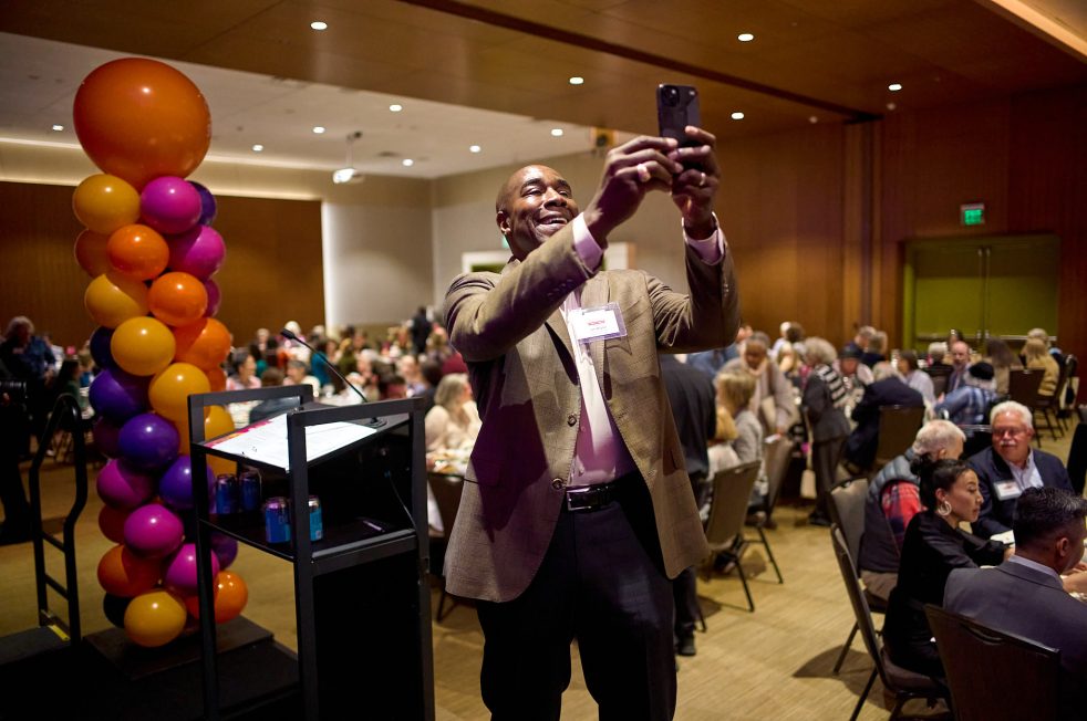 Man smiling while taking a photo on his phone at a community fundraiser supporting senior services in Seattle