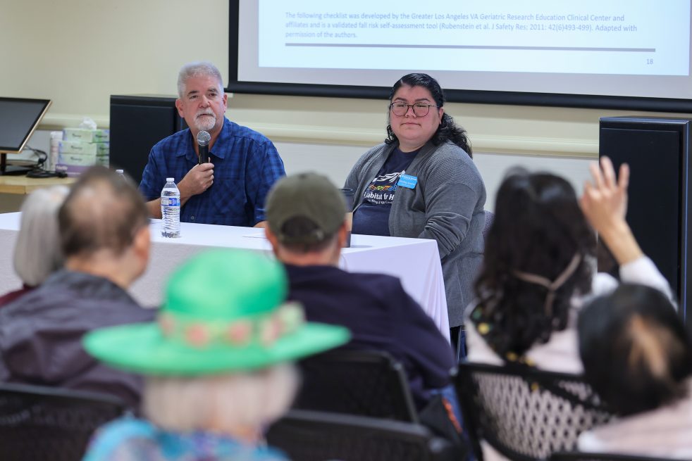 photo of two people seated at a table at the front of a meeting room. One person holding a microphone. Approximately 6 people in the audience are seen from behind. One person is raising their hand.