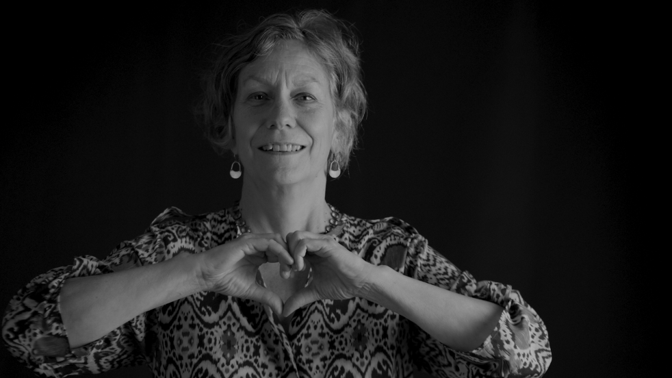 A smiling older woman forms a heart shape with her hands against a dark backdrop to highlight Sound Generations' Elder Education Institute and their commitment to caregiver workshops, education for seniors, and aging care services in Seattle and King County.